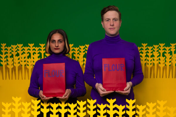 Two individuals wearing purple turtle necks, holding bags of flour