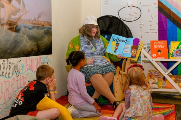 A woman in a tabard an white hat sits on a char and reads a story book to a group of children.