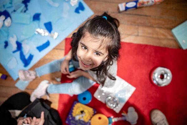 A young girl looks up to the camer smiling. She is sat on the floor surrounded by arts and crafts