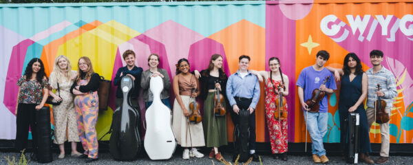 A group of smiling musicians stand in a row in front of a colourfully painted background.